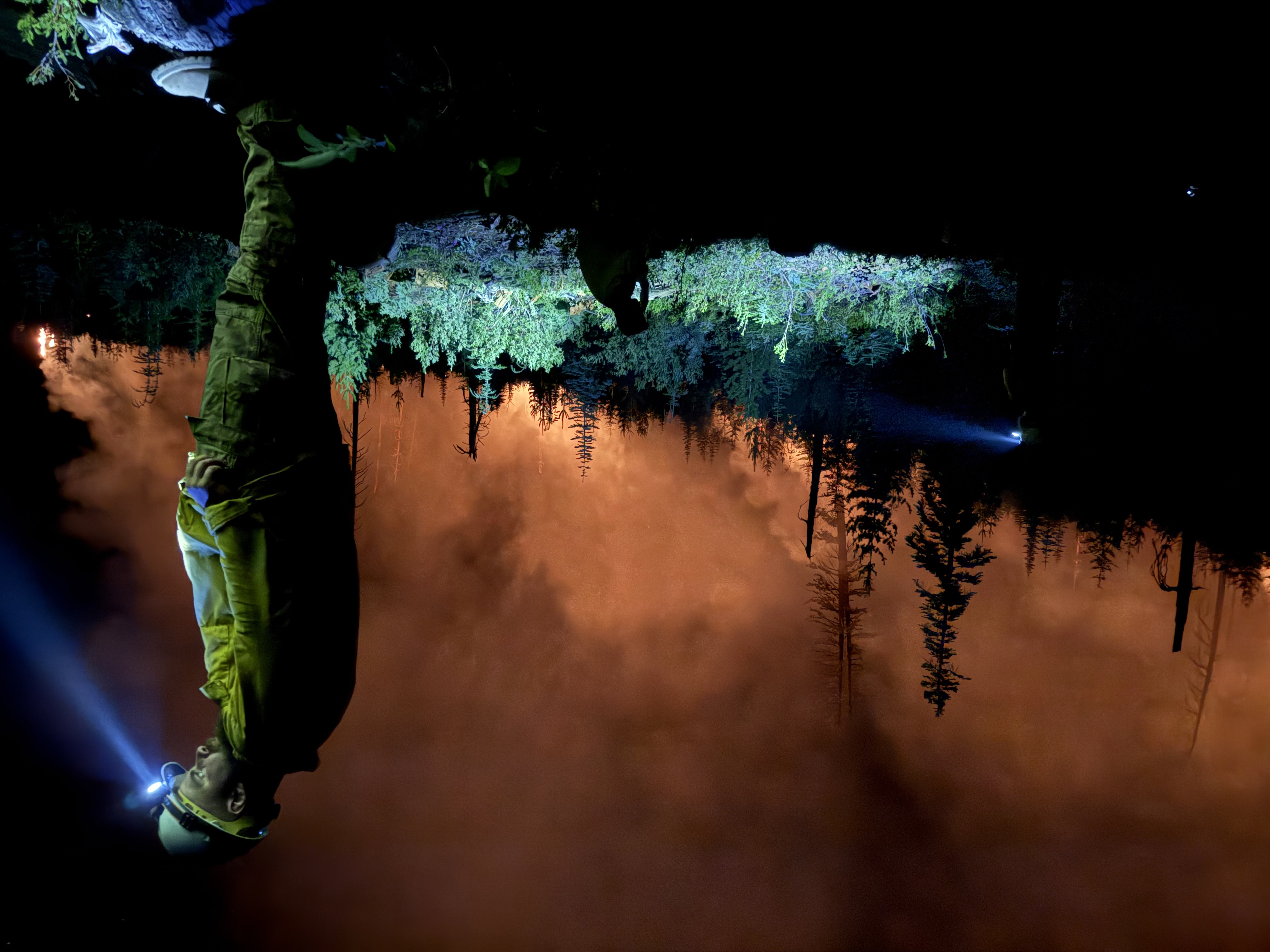 Night operations on the fireline, with wildland firefighters working by headlamp as active fire burns through timber behind them.