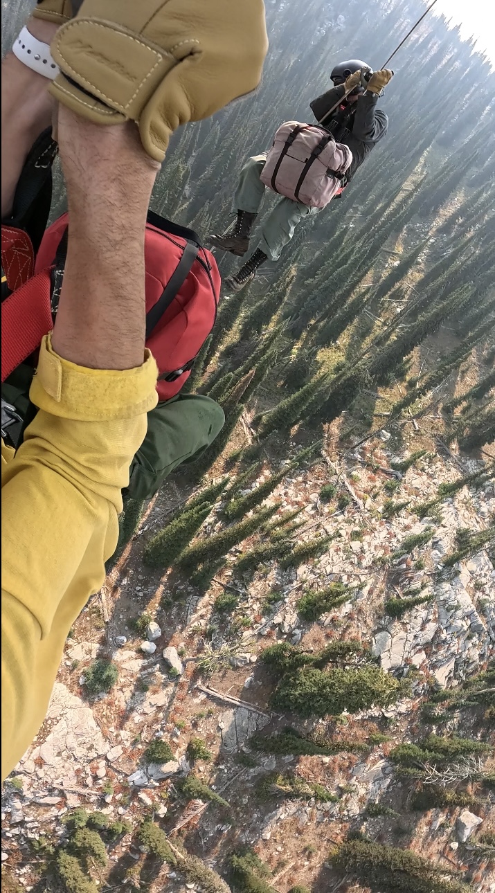 Two heli-rappellers descend side by side over rugged mountain terrain, carrying the gear needed to begin work after insertion.