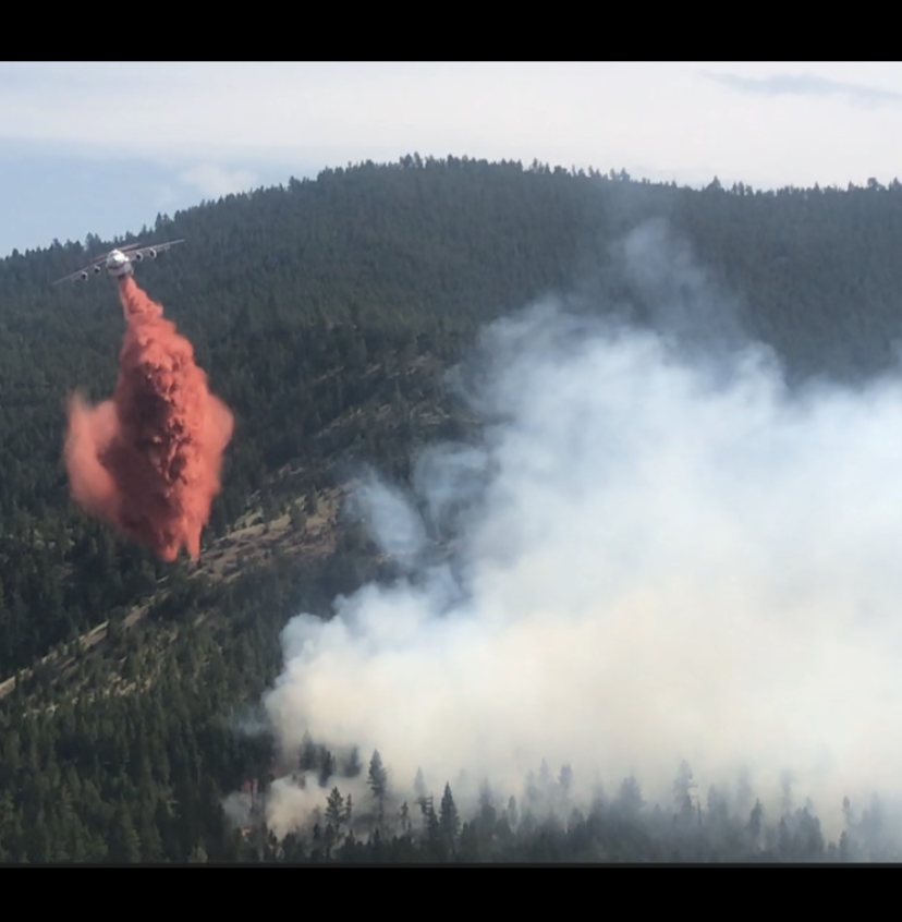 An air tanker drops retardant along a wildfire’s edge, supporting rappellers working below in rugged, forested terrain.
