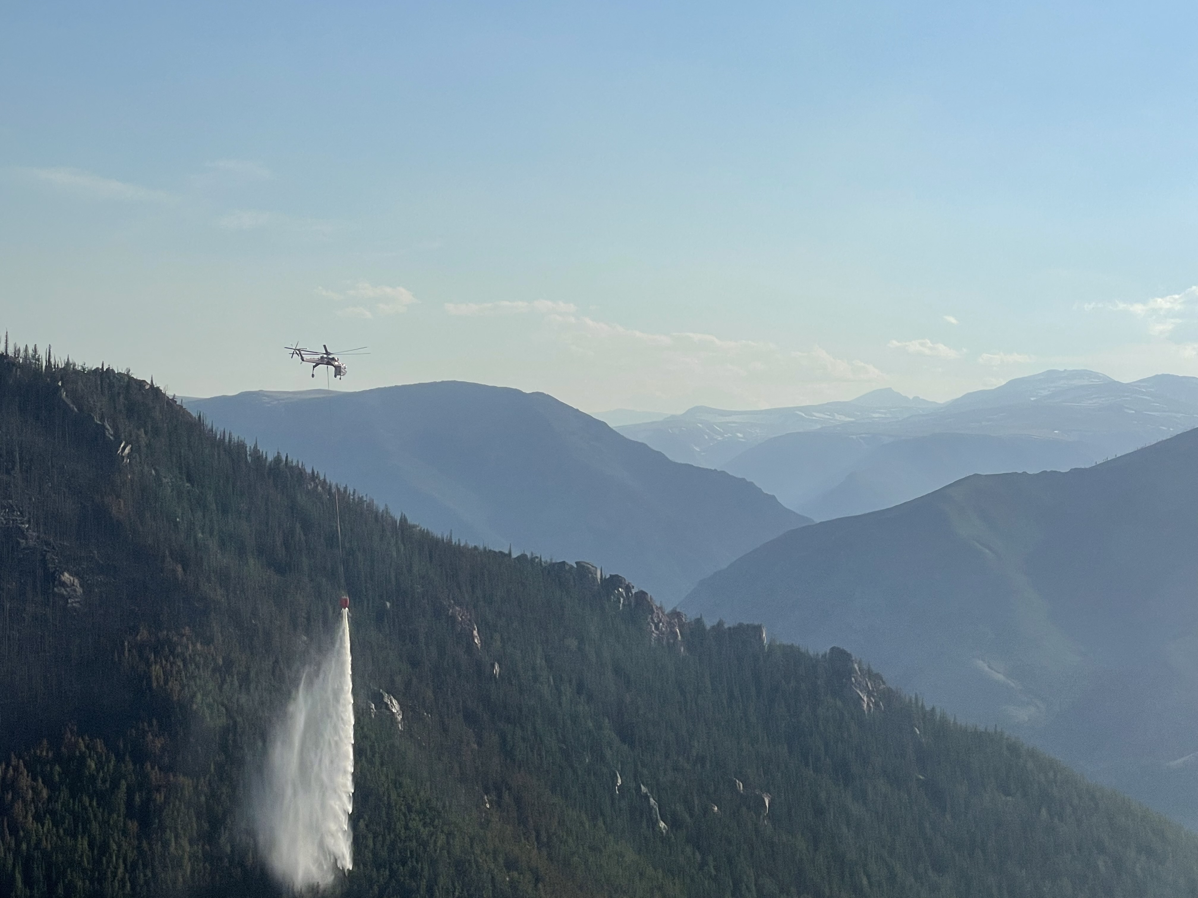 Helicopter bucket drops support wildfire suppression in steep mountain terrain, where aviation resources and rappellers can reach fires with limited ground access.