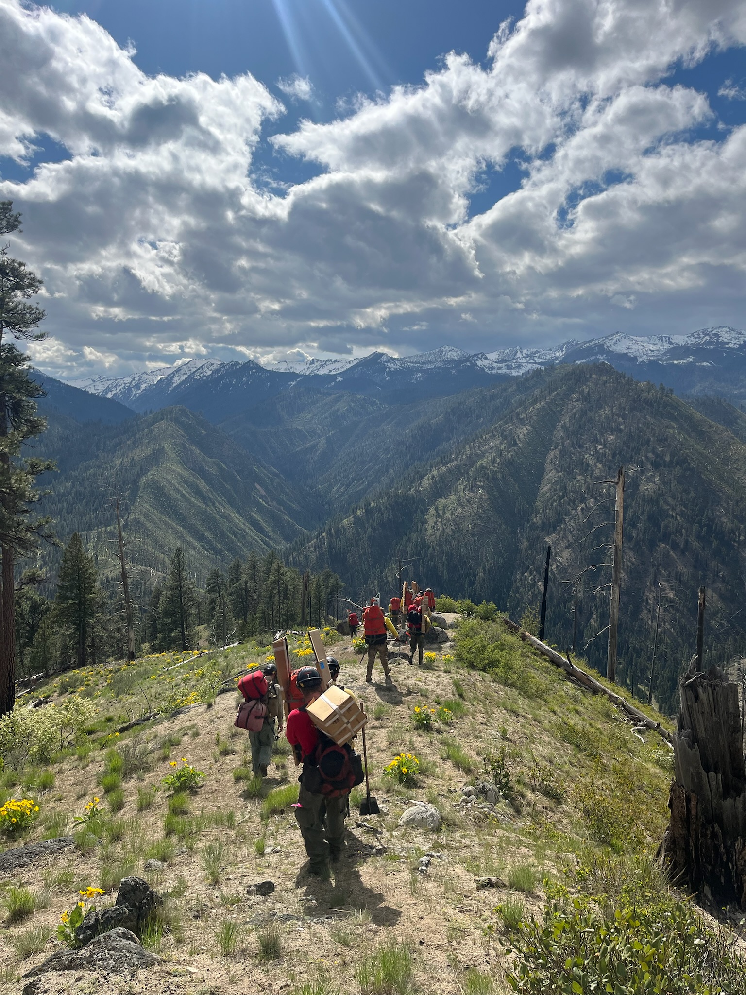 Heli-rappellers pack gear across a high mountain ridge, showing the demanding backcountry travel that comes with remote wildland fire assignments.
