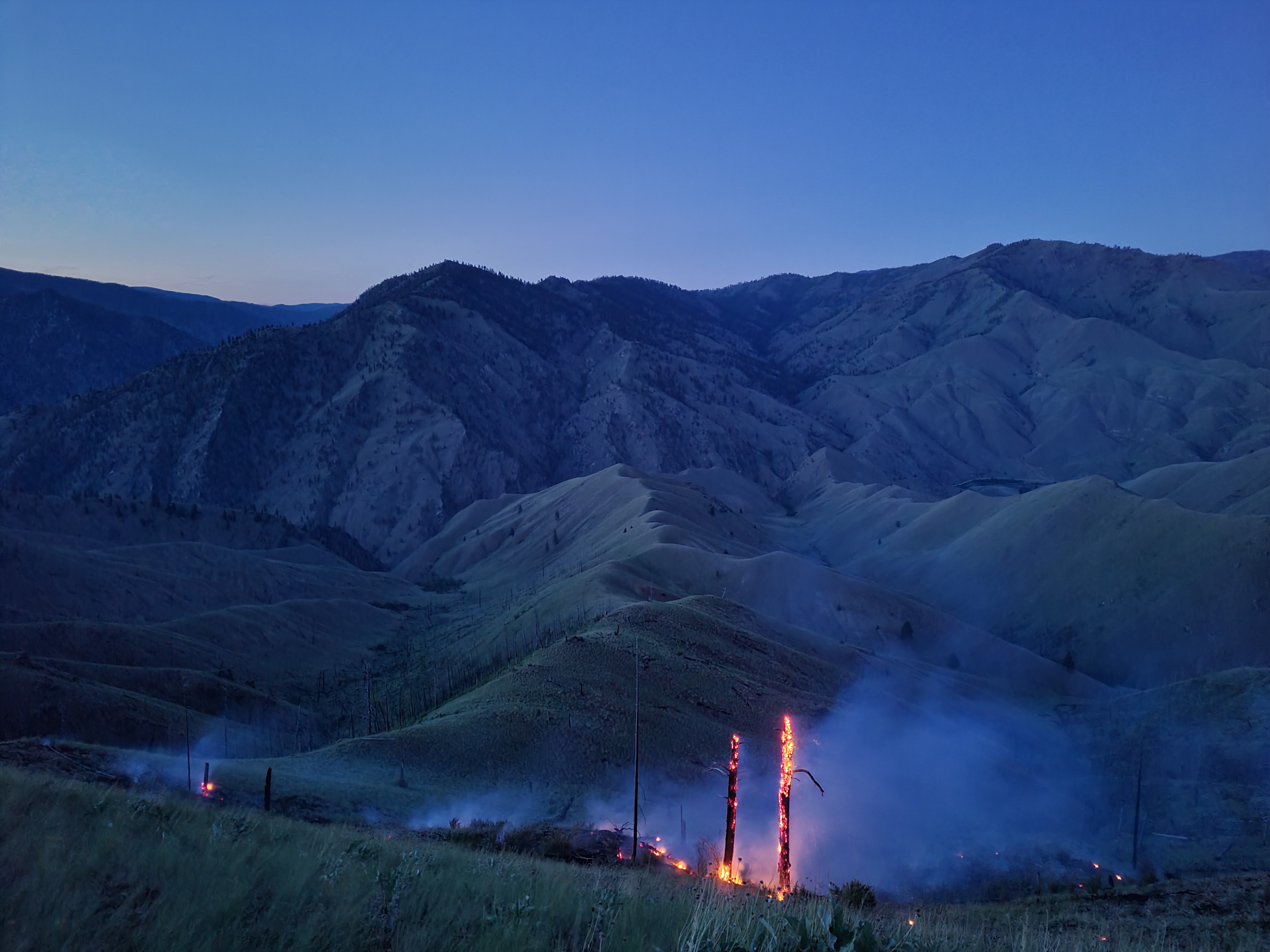 Dusk settles over a remote wildfire as scattered heat and burning snags remain active across steep mountain terrain.