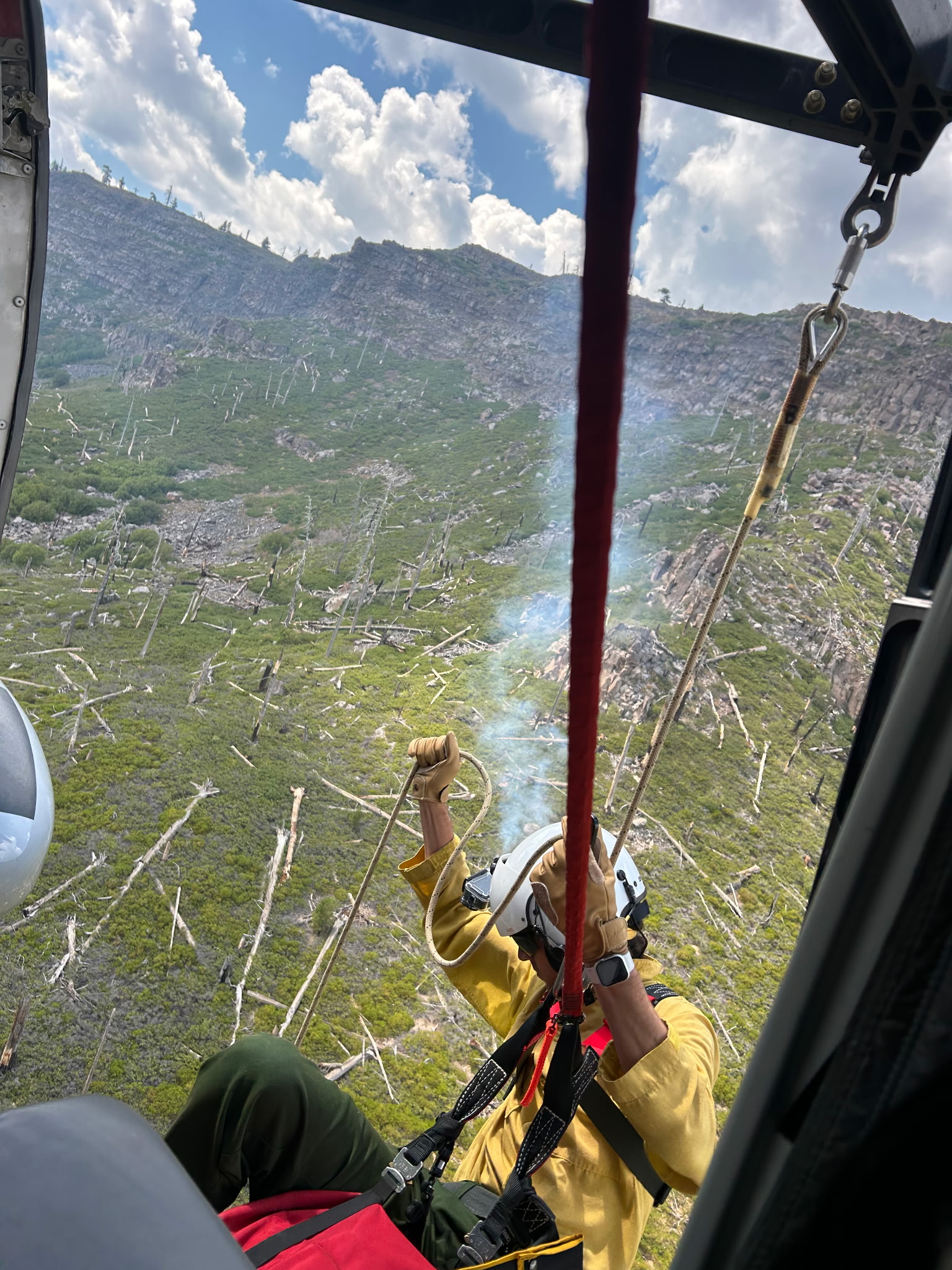 A heli-rappeller prepares to descend toward a remote wildfire, with smoke and steep terrain visible below the aircraft.