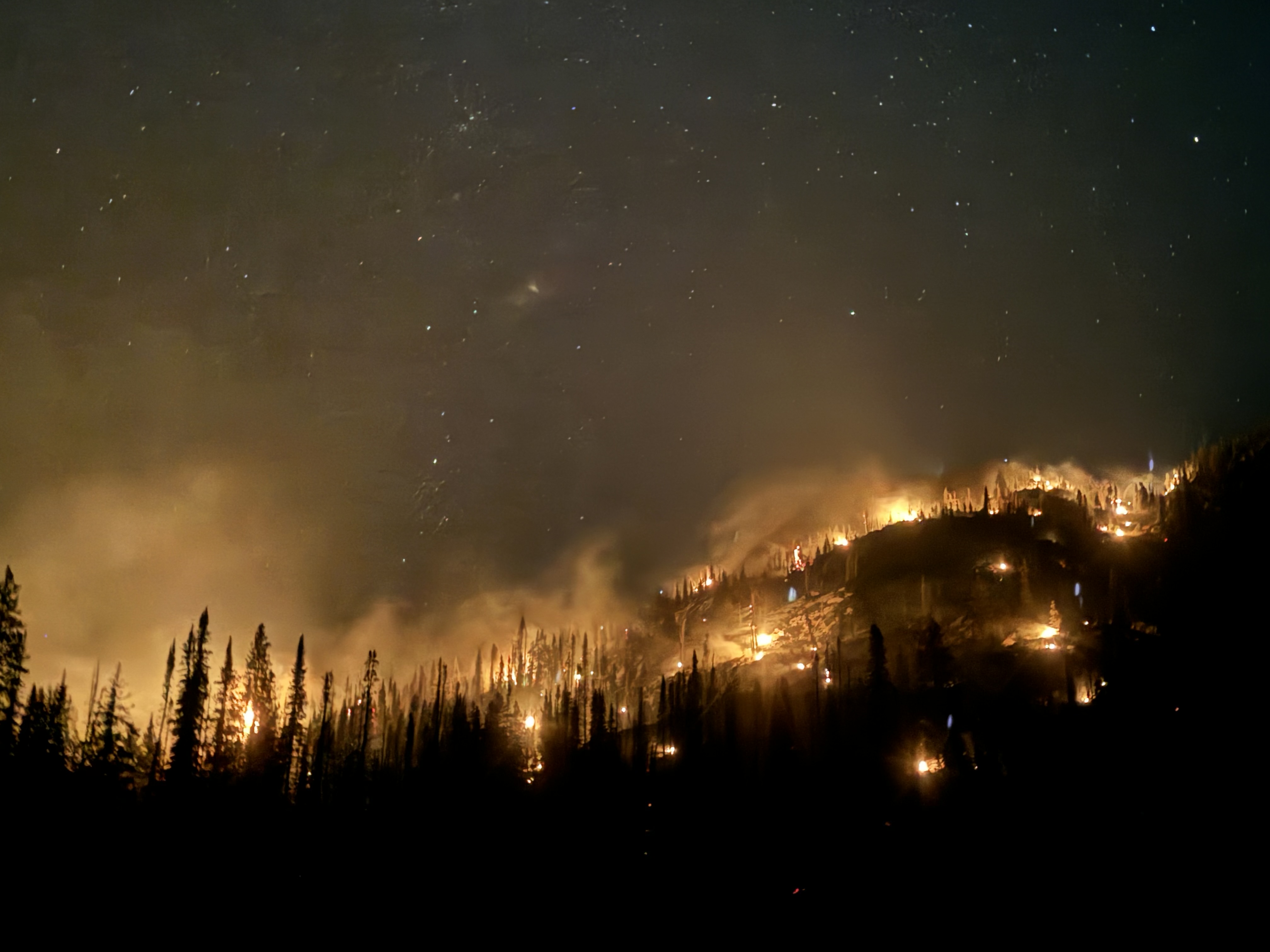 Wildfire spreads across a mountain ridge beneath the night sky, illuminating the remote terrain where crews work through extended fire operations.